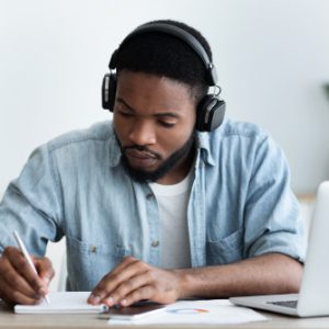 A young Black man with headphones on writing on a pad of paper while sitting at a desk.